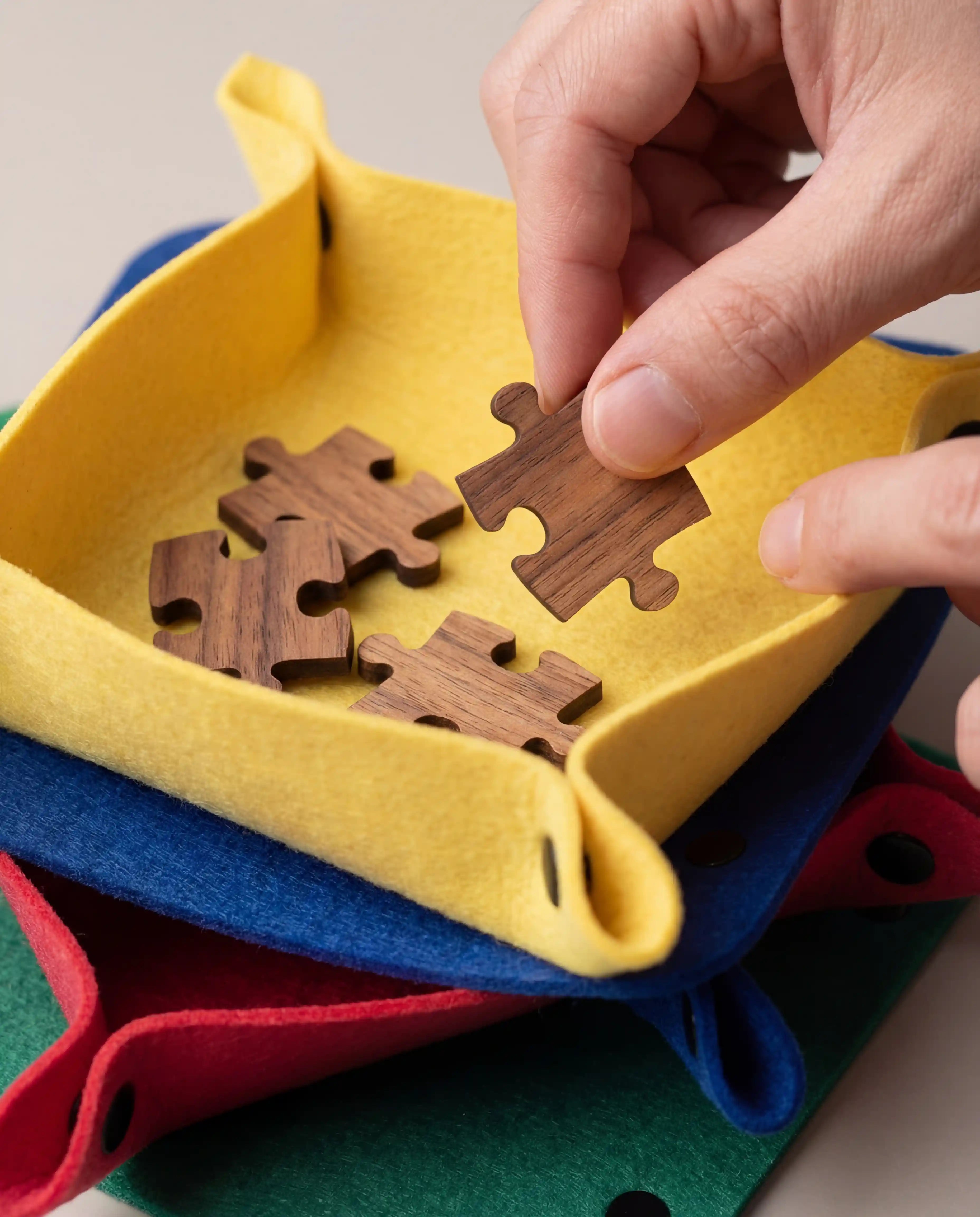 Gros plan sur une main manipulant une pièce de puzzle en bois de tilleul dans un plateau de tri en feutre jaune, illustrant la protection des textures et le confort d'assemblage.